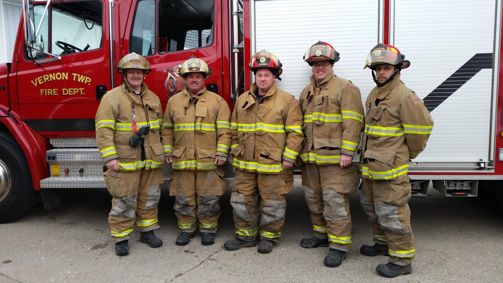 Five firefighters dressed in full firefighting gear while standing outside in front of a fire engine.