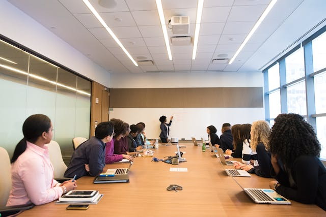 A large group of people sitting around a long rectangle table while a woman writes on a dry erase board while teaching them.